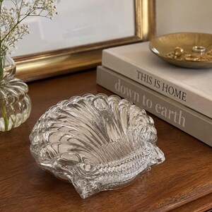 May include: A clear glass dish shaped like a scalloped seashell with a ribbed texture and a glossy finish. The object features a wide, fan-like design with curved edges and a central hinge-like detail at the base, reflecting light on its surface.