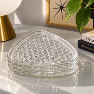May include: A stack of four clear glass plates with a rounded triangular shape. Each plate features a textured, geometric diamond pattern on the surface, creating a faceted appearance. The edges are smooth and transparent, reflecting light.