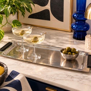 May include: A rectangular stainless steel serving tray with black handles on each end. The tray holds two ribbed glass coupes filled with clear liquid and green olives on gold picks, and a small stainless steel bowl filled with green olives.