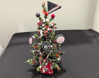Puede incluir: Un pequeño árbol de hoja perenne artificial decorado con adornos temáticos de la Universidad de Georgia, que incluyen discos redondos de fútbol americano, un banderín negro con el texto GEORGIA, cuentas rojas y negras, y una base roja con letras blancas.