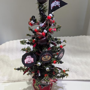 May include: A miniature Christmas tree decorated with Ohio State University Buckeyes themed ornaments. The tree features a black flag with "OHIO STATE" text, red and white accents, and a base with "BUC" text.