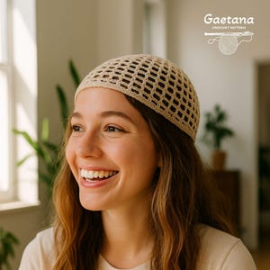 Puede incluir: Un gorro de ganchillo beige con un diseño de tejido abierto. La persona que lo lleva tiene el pelo largo y castaño. El texto "Gaetana Crochet Pattern" es visible. El gorro parece hecho a mano, un accesorio de verano.