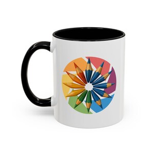 Può includere: Tazza in ceramica bianca con manico e interno neri. La tazza presenta un design colorato di matite disposte a cerchio, con uno sfondo sfumato arcobaleno. Le matite sono nelle tonalità dell'arancione, del giallo, del verde e del blu.