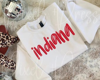 May include: A white sweatshirt with the word "Indiana" in red, cursive lettering. The sweatshirt is displayed on a fluffy, light-coloured surface, with a small disco ball and a leopard print accessory visible.