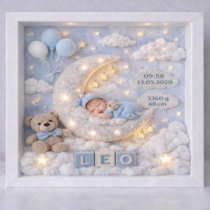Puede incluir: Recuerdo de bebé enmarcado con un bebé durmiendo en una luna creciente, rodeado de nubes, estrellas y globos. Incluye un osito de peluche, el nombre "LEO" y detalles de nacimiento: 3360 g y 48 cm.