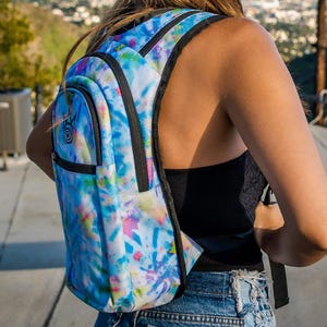 Op de afbeelding: Een tie-dye rugzak met een levendige mix van blauw, roze en gele tinten. De rugzak heeft zwarte randen, een voorvak en verstelbare banden. Het ontwerp bevat een spiraalsymbool op het voorvak. Geschikt voor school of reizen.