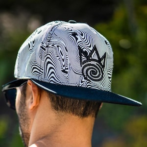 May include: A black and white patterned baseball cap with a flat brim. The cap features a swirling, wavy design and a black embroidered emblem on the front. The brim is solid black.