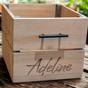 May include: A light-colored wooden storage crate with a black handle. The name "Adeline" is carved into the front. The crate is sitting on a wooden surface, with plants in the background.