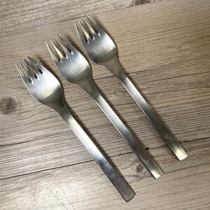 May include: Three silver-colored metal forks arranged diagonally. Each fork features a four-tine head and a flat, straight handle with a smooth, brushed finish. The design is simple and uniform, showcasing a metallic sheen across the surface.