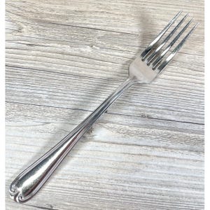 May include: A stainless steel fork with four tines and a long, slender handle. The handle has a decorative, curved design at the base. The fork is designed for eating and is likely part of a dining set.
