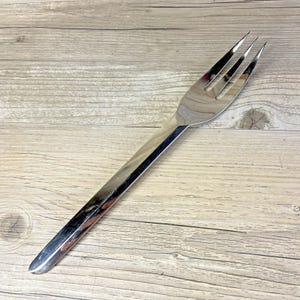 Op de afbeelding: Een glanzende roestvrijstalen vork met drie tanden. De vork heeft een lange, slanke steel en is ontworpen om te serveren. Het bestek ligt op een lichtbruin houten oppervlak.