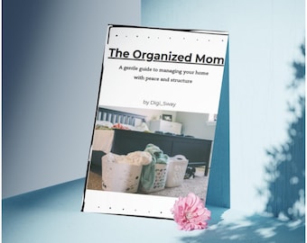 May include: A book titled "The Organized Mom" with the subtitle "A gentle guide to managing your home with peace and structure" by Digi_Sway. The book is propped up against a blue surface with a pink flower and shadows.