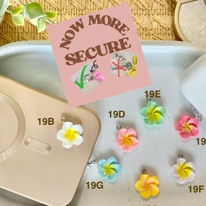 May include: A collection of colorful flower-shaped charms, including white, yellow, pink, blue, and green, with silver-tone metal findings. The charms are displayed with a phone and a white case. The text "NOW MORE SECURE" is visible.