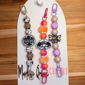 May include: Three beaded lanyards with metal clips. The left features brown and patterned beads with a round charm reading Dragons and Romance. The center has orange and pink beads with a Bat Boys Fan Club charm. The right has purple beads and a Faerie Smut book charm.