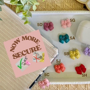 May include: A collection of colorful bow charms in various shades, including pink, teal, yellow, and red, displayed on a white oval tray. A white AirPod case is also present. A pink square with the text "NOW MORE SECURE" is in the image.