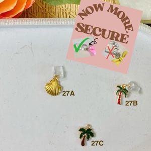 Op de afbeelding: Een selectie van kleine, decoratieve bedels, waaronder een gouden zeeschelp, een palmboom en een ander palmboomontwerp. De bedels zijn bevestigd aan doorzichtige oorhaakjes. Een roze bord leest "NOW MORE SECURE".