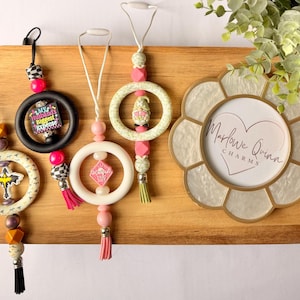 Puede incluir: Una bandeja de madera contiene varios ambientadores para coche y un marco decorativo. Los ambientadores presentan varios diseños, incluyendo una cruz, un anillo con el texto "My Husband's Biggest Expense" y un diamante rosa. El marco tiene el texto "Marlowe Quinn CHARMS."