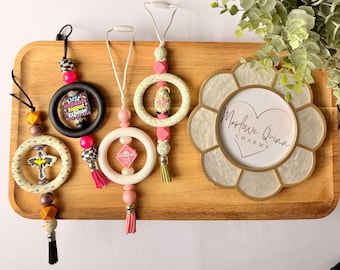 May include: A wooden tray holds several car air fresheners and a decorative frame. The air fresheners feature various designs, including a cross, a ring with the text "My Husband's Biggest Expense," and a pink diamond. The frame has the text "Marlowe Quinn CHARMS."
