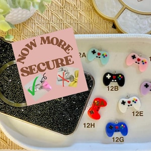 May include: A black bedazzled phone case with a magnetic ring, a pink card with the text "NOW MORE SECURE", and colorful game controller charms in various colors. The charms are in light blue, black, red, white, blue, pink, and purple.