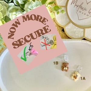 May include: The image showcases two small teddy bear charms, one light brown and one beige, hanging from a clear clasp. A pink sign reads "NOW MORE SECURE" with a comparison of secure and insecure clasping methods. The background includes greenery and a decorative tray.