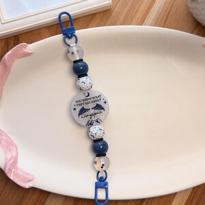 May include: A beaded lanyard featuring a central circular charm with silver glitter, a crescent moon, bat wings, and the text "YOU KNOW WHAT THEY SAY ABOUT wingspan". The design includes blue and white star-patterned beads, blue spheres, and blue clips.