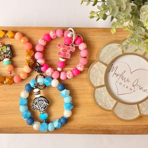 May include: Three beaded wristlet keychains in shades of pink, orange, and blue, each with a charm. One keychain has the text "Fresh Out of Fucks". A decorative flower-shaped coaster with the text "Marlowe Quinn Charms" is also present.