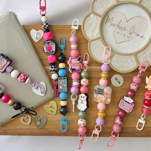 Könnte beinhalten: Eine Sammlung von Perlen-Telefonanhängern in Rosa, Schwarz, Weiß und Lila. Die Anhänger zeigen Herz-, Buch- und Engel-Designs mit den Texten "fueled by coffee & books" und "you are amazing".