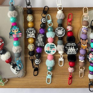 Puede incluir: Una colección de llaveros de cuentas con varios dijes. Los llaveros presentan una mezcla de cuentas coloridas, acentos esponjosos y dijes decorativos con imágenes y texto. Cada llavero tiene un cierre de metal.
