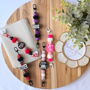 May include: A collection of colorful beaded keychains with various charms, including text that reads "good girl" and "I prefer my fairytales dark." The keychains feature black, red, pink, and purple beads, along with fluffy accents and silver-tone hardware.