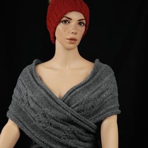 Op de afbeelding: Een rode gebreide muts met een imitatiebont pompon en een grijze gebreide omslagdoek. De muts is dieprood van kleur en de omslagdoek is middengrijs. De omslagdoek is ontworpen om over de schouders te worden gedragen.