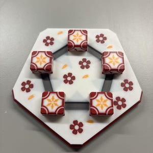 May include: A white pentagonal board featuring a dark gray frame and red flower patterns with scattered orange petals. Five square red blocks with a central yellow star design are positioned on the board, each resting within the dark gray frame.