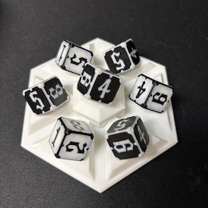 May include: A set of six black and white six-sided dice arranged on a white hexagonal base. Each die features high-contrast black faces with white numbers in a decorative font, displaying various numerical values including one, two, four, five, and six.
