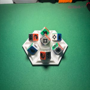 May include: A white hexagonal dice tray holding seven multi-sided dice. The dice are white with various symbols in black, orange, green, and blue. The tray sits on a green surface.