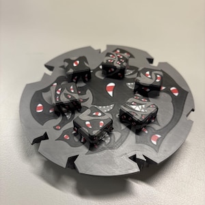 Puede incluir: Un tablero de juego circular gris con bordes dentados que presenta un patrón gráfico en negro, gris y rojo de múltiples ojos y dientes. Cinco dados de juego cuadrados a juego están dispuestos sobre la superficie, cada uno con el mismo motivo.