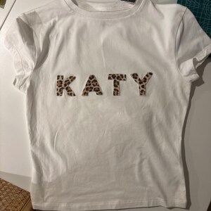 Puede incluir: Una camiseta blanca con el nombre "KATY" en letras con estampado de leopardo en el pecho. La camiseta tiene mangas cortas y cuello redondo. La tela parece ser suave y ligera.