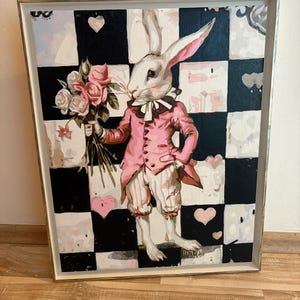 May include: A framed painting depicts a rabbit dressed in a pink suit and holding a bouquet of roses. The rabbit stands on a black and white checkered background with pink hearts. The artwork has a silver frame.