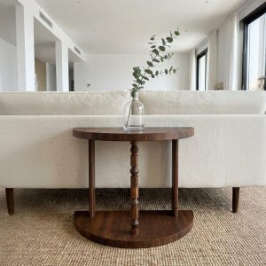 Puede incluir: Una mesa auxiliar semicircular de color marrón oscuro con un soporte central decorativo y dos patas. Un jarrón de cristal transparente con follaje verde está sobre la mesa. La mesa está frente a un sofá color crema.