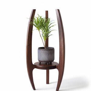 May include: A dark brown wooden plant stand with a modern design. The stand features a circular shelf and a tall, vertical support. A gray, ribbed ceramic pot with a green plant sits on the shelf.