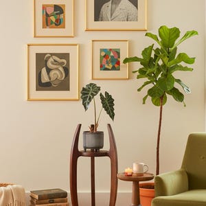 May include: A living room scene featuring framed artwork, a potted plant on a wooden stand, and a green armchair. The artwork includes a portrait and abstract pieces. A small side table holds a mug and candles.