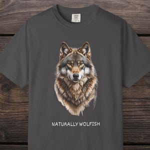 Op de afbeelding: Donkergrijs t-shirt met een gedetailleerde illustratie van een wolfskop. De wolf heeft bruin, grijs en wit haar. De tekst "NATURALLY WOLFISH" is wit onder de wolfafbeelding gedrukt. Het shirt wordt gepresenteerd op een houten oppervlak.
