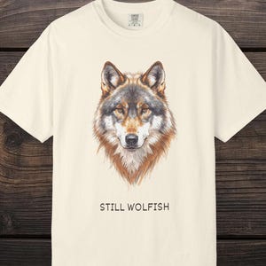 Op de afbeelding: Crèmekleurig T-shirt met een gedetailleerde illustratie van een wolfskop in bruin, grijs en wit. De tekst "STILL WOLFISH" staat onder de afbeelding. Het shirt heeft een klassieke ronde hals en korte mouwen.