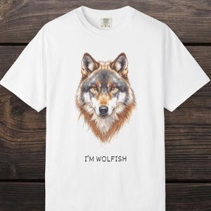 Op de afbeelding: Wit t-shirt met een gedetailleerde illustratie van een wolfskop in bruin, grijs en beige. De tekst "I'M WOLFISH" is onder de afbeelding gedrukt. Het shirt is gemaakt van een zacht materiaal.