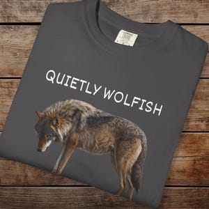 Op de afbeelding: Donkergrijs t-shirt met de witte tekst "QUIETLY WOLFISH". Het shirt toont een gedetailleerde illustratie van een wolf in bruine en grijze tinten.