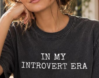 May include: A charcoal gray short-sleeve t-shirt with a crew neckline. The shirt features the white text "IN MY INTROVERT ERA" printed in a distressed, typewriter-style font across the center of the chest. The fabric has a faded, vintage wash.