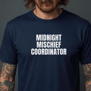 Peut inclure: T-shirt bleu marine avec l'inscription blanche "MIDNIGHT MISCHIEF COORDINATOR". Le t-shirt est de style col rond et semble être fait d'un tissu doux et respirant. La personne qui porte le t-shirt a des tatouages sur les bras.