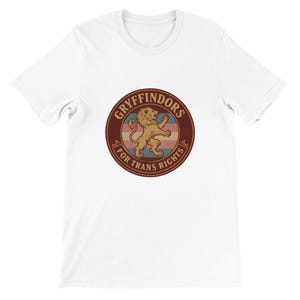 Pode incluir: Camiseta branca com um design circular com um leão e as palavras "Gryffindors For Trans Rights". O design usa tons de marrom, vermelho e azul. A camiseta é feita de tecido macio.