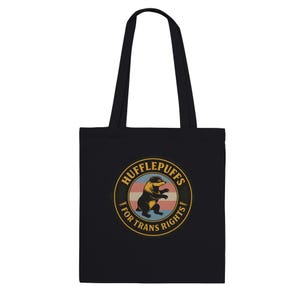 Pode incluir: Uma sacola preta com um gráfico circular apresentando um texugo no centro. O texto "HUFFLEPUFFS FOR TRANS RIGHTS" está impresso em amarelo ao redor do texugo. O fundo do círculo consiste em listras horizontais azul-claras, rosas e brancas.