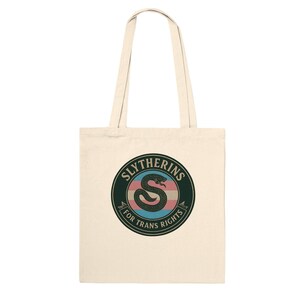 Pode incluir: Uma sacola de lona cor de creme com um gráfico circular de borda verde-escura. Dentro do círculo, há uma cobra preta enrolada sobre um fundo listrado em azul claro, rosa e branco, com o texto SLYTHERINS FOR TRANS RIGHTS impresso.