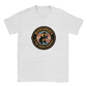 May include: A light gray short-sleeve t-shirt featuring a circular graphic on the chest. The design includes a black badger illustration in the center, surrounded by the text "HUFFLEPUFFS FOR TRANS RIGHTS" against a background of horizontal stripes.