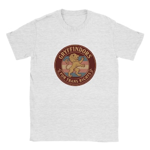 Può includere: Una t-shirt grigio chiaro mélange con una grafica circolare sul petto. Il design presenta un leone dorato al centro, circondato da strisce orizzontali rosa, bianche e blu, con il testo GRYFFINDORS FOR TRANS RIGHTS.